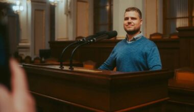 Mężczyzna w niebieskim swetrze stoi za mównicą i uśmiecha się, przemawiając publicznie.