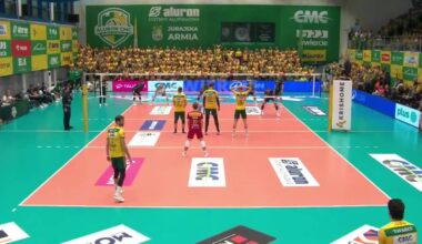 Mecz siatkówki z graczami na boisku i kibicami na trybunach.