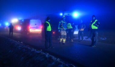 Strażacy w kamizelkach odblaskowych i pojazdy ratunkowe z włączonymi niebieskimi światłami na tle nocnego krajobrazu.