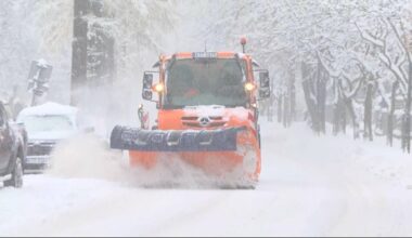 Pomarańczowy pług śnieżny z łyżką odśnieżającą na zaśnieżonej drodze podczas opadów śniegu.