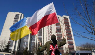 Osoba trzymająca polską i ukraińską flagę na tle budynku mieszkalnego.