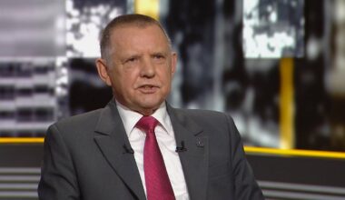 Marian Banaś w garniturze z czerwonym krawatem, siedzący i udzielający wywiadu.