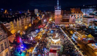 Nocny widok na zatłoczony jarmark bożonarodzeniowy w historycznym centrum miasta, z licznymi straganami, iluminacjami i budynkami.