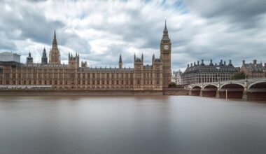 Szeroki kadr ukazujący Pałac Westminsterski w Londynie z wieżą zegarową, nad rzeką Tamizą, przy zachmurzonym niebie.