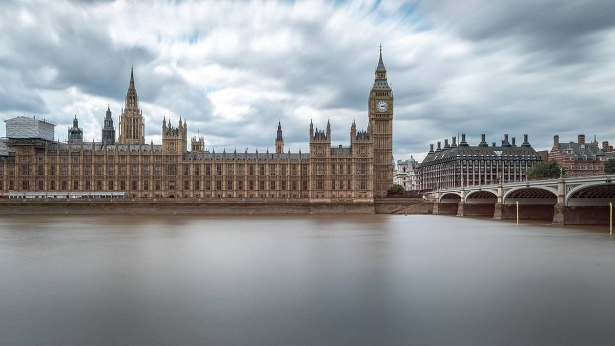Szeroki kadr ukazujący Pałac Westminsterski w Londynie z wieżą zegarową, nad rzeką Tamizą, przy zachmurzonym niebie.