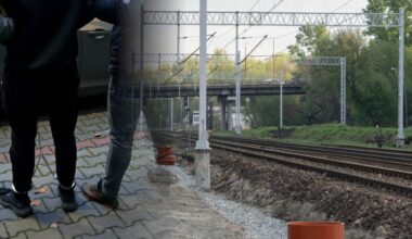 Fragment sylwetek dwóch osób stojących na chodniku obok torowiska kolejowego.