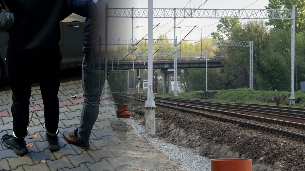 Fragment sylwetek dwóch osób stojących na chodniku obok torowiska kolejowego.