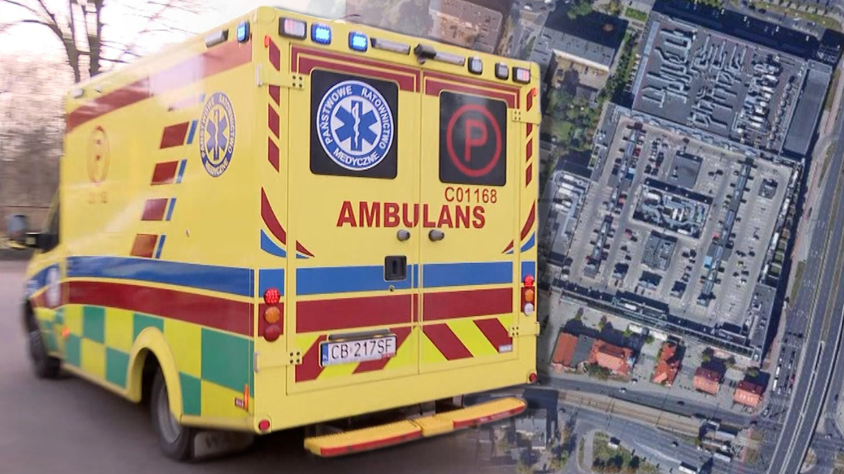 Karetka pogotowia z napisem "AMBULANS" na tle widoku z lotu ptaka na parking galerii handlowej.
