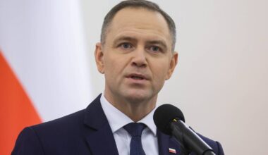 Mężczyzna w ciemnym garniturze i białej koszuli z krawatem stoi przy mównicy, przemawiając do mikrofonu. W tle widoczna jest polska flaga.