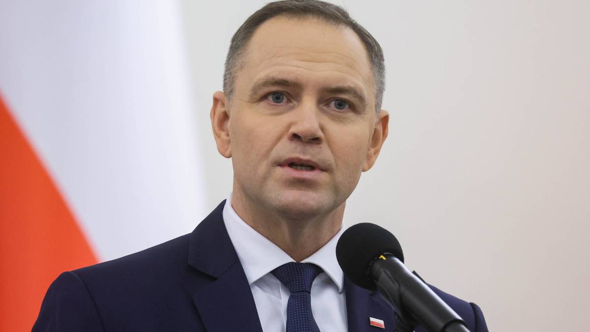 Mężczyzna w ciemnym garniturze i białej koszuli z krawatem stoi przy mównicy, przemawiając do mikrofonu. W tle widoczna jest polska flaga.