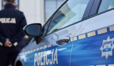 Fragment radiowozu policyjnego z napisem POLICJA i symbolem policji. W tle stoi policjant w mundurze.