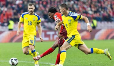 Trzech piłkarzy na boisku piłkarskim, dwóch w żółtych strojach i jeden w czerwonym, rywalizuje o piłkę.