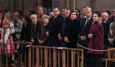 Grupa osób w ciemnych ubraniach stoi w rzędzie, patrząc w tym samym kierunku. Na pierwszym planie widoczne są drewniane barierki.