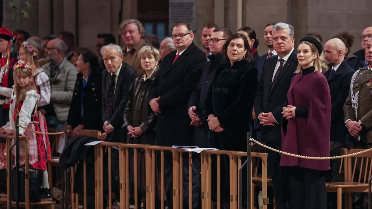 Grupa osób w ciemnych ubraniach stoi w rzędzie, patrząc w tym samym kierunku. Na pierwszym planie widoczne są drewniane barierki.