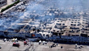 Widok z lotu ptaka na spalony dach hali handlowej, z którego unosi się dym. Na parkingu przed budynkiem widoczne są samochody i pojazdy strażackie.