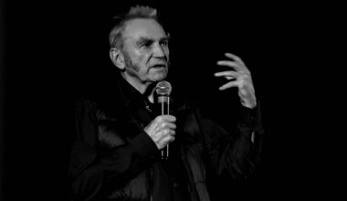 Starszy mężczyzna w garniturze trzyma mikrofon i gestykuluje prawą ręką podczas wystąpienia na czarnym tle.