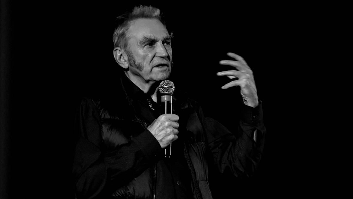 Starszy mężczyzna w garniturze trzyma mikrofon i gestykuluje prawą ręką podczas wystąpienia na czarnym tle.