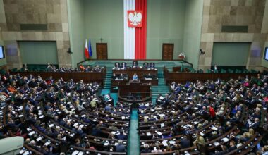 Widok z góry na salę plenarną Sejmu Rzeczypospolitej Polskiej, z posłami siedzącymi w ławach i flagą Polski w tle.