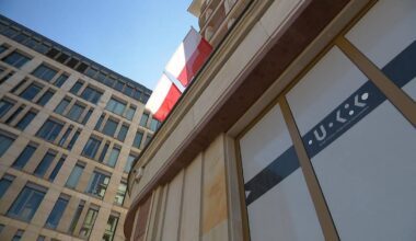 Fragment budynku z logo UOKiK i polską flagą na tle błękitnego nieba.