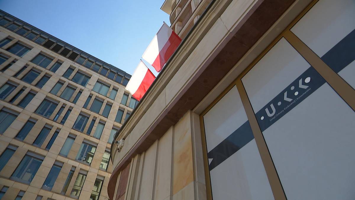 Fragment budynku z logo UOKiK i polską flagą na tle błękitnego nieba.
