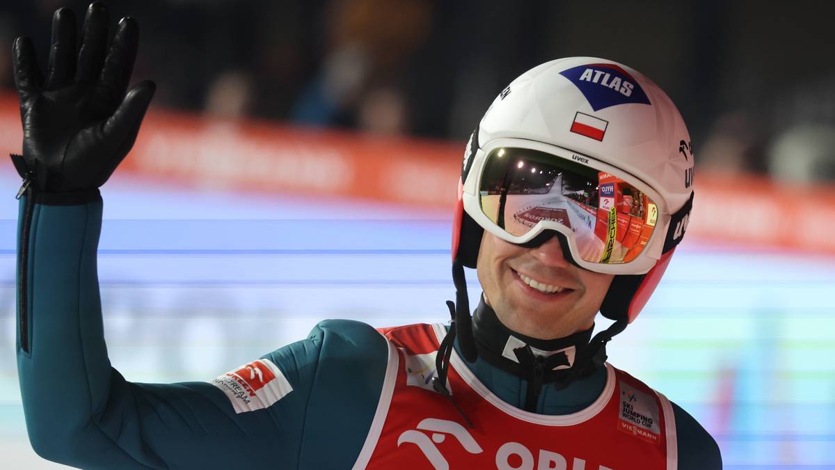 Kamil Stoch w kasku i okularach narciarskich pozdrawia, machając ręką.