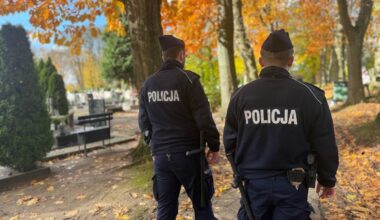 Dwóch policjantów w mundurach idzie przez alejkę z drzewami i liśćmi na ziemi.