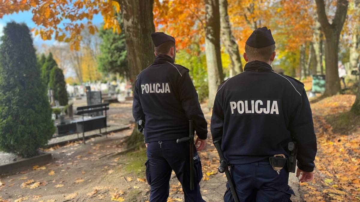 Dwóch policjantów w mundurach idzie przez alejkę z drzewami i liśćmi na ziemi.