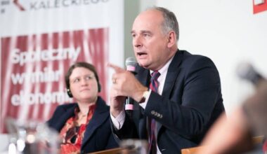 Mężczyzna w garniturze przemawia do mikrofonu, wskazując palcem, obok siedzi kobieta w słuchawkach.