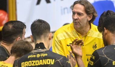 Trener w żółtej koszulce sportowej przemawia do grupy zawodników siatkówki w czarnych strojach, skupionych wokół niego.