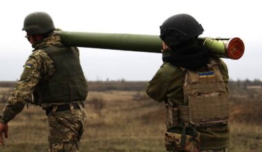 Dwóch żołnierzy w mundurach taktycznych i hełmach niesie na ramieniu długi, zielony pocisk.