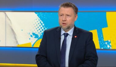 Mężczyzna w ciemnym garniturze i krawacie, siedzący przed kolorowym tłem studyjnym.