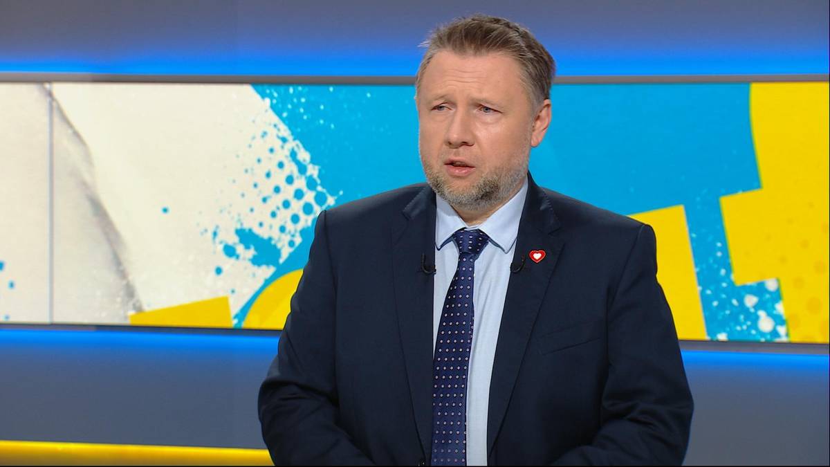 Mężczyzna w ciemnym garniturze i krawacie, siedzący przed kolorowym tłem studyjnym.