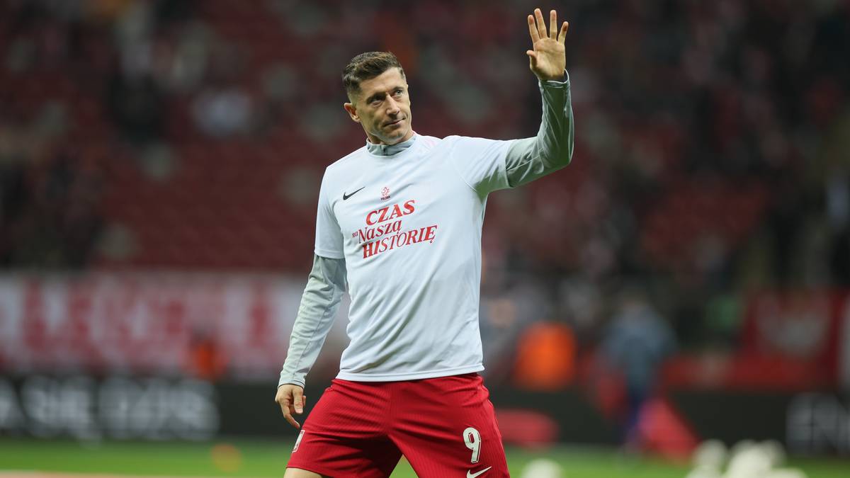 Robert Lewandowski w białej koszulce reprezentacji Polski i czerwonych spodenkach z numerem 9 na boisku, macha ręką do publiczności.