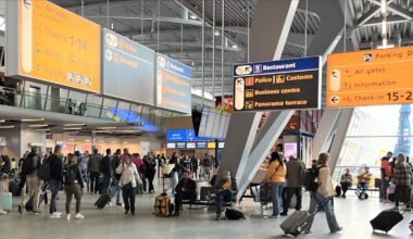 Grupa ludzi z bagażami porusza się po jasnym, przestronnym wnętrzu lotniska, obok schodów ruchomych i pod wiszącymi tablicami informacyjnymi.