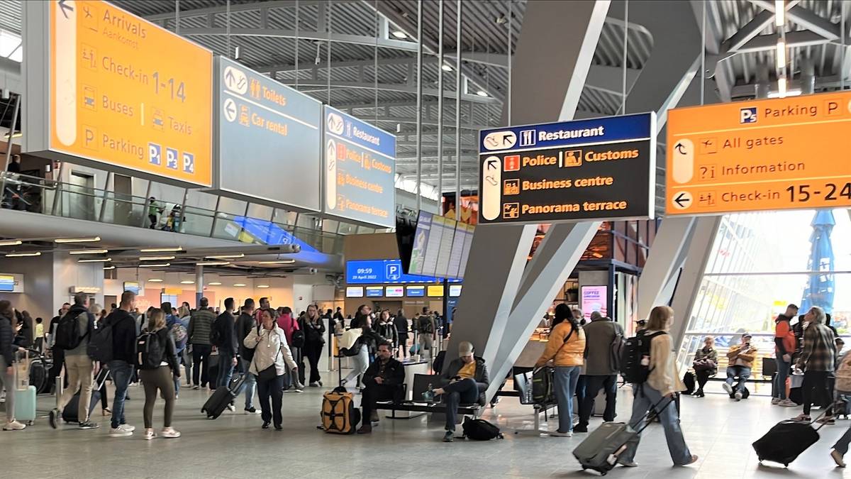 Grupa ludzi z bagażami porusza się po jasnym, przestronnym wnętrzu lotniska, obok schodów ruchomych i pod wiszącymi tablicami informacyjnymi.