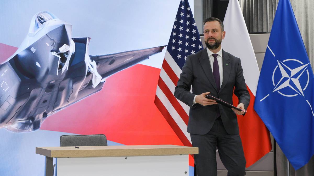 Szef MON Mariusz Błaszczak stoi obok flag Polski, USA i NATO, z sylwetką samolotu F-35 w tle.