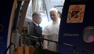 Papież Leon XIV w białej szacie wchodzi po schodach samolotu, obok niego stoi mężczyzna w garniturze.