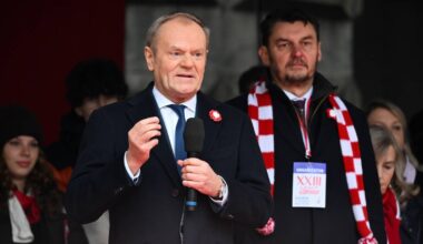 Premier Donald Tusk przemawia na tle grupy osób, trzymając mikrofon.