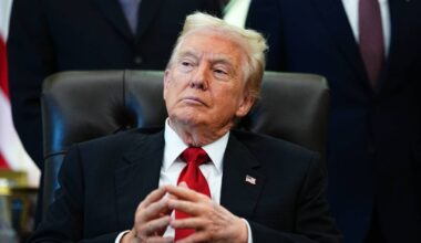 Donald Trump siedzący za biurkiem w garniturze, z dłońmi złożonymi przed sobą.