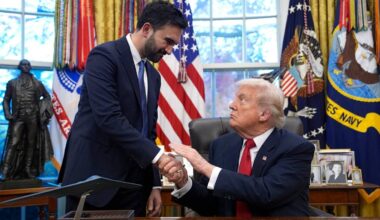 Donald Trump i Zohran Mamdani ściskają sobie dłonie w Gabinecie Owalnym Białego Domu, siedząc przy biurku. W tle widoczne są flagi Stanów Zjednoczonych.