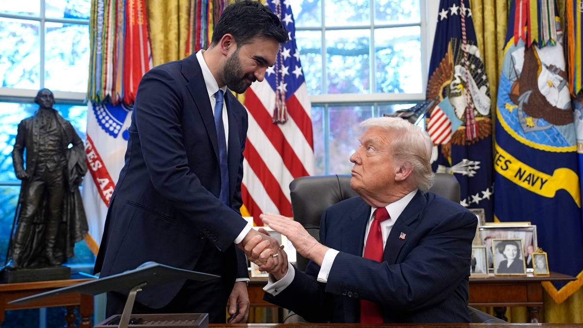 Donald Trump i Zohran Mamdani ściskają sobie dłonie w Gabinecie Owalnym Białego Domu, siedząc przy biurku. W tle widoczne są flagi Stanów Zjednoczonych.