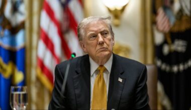 Donald Trump w garniturze i żółtym krawacie, siedzący przed flagami USA.