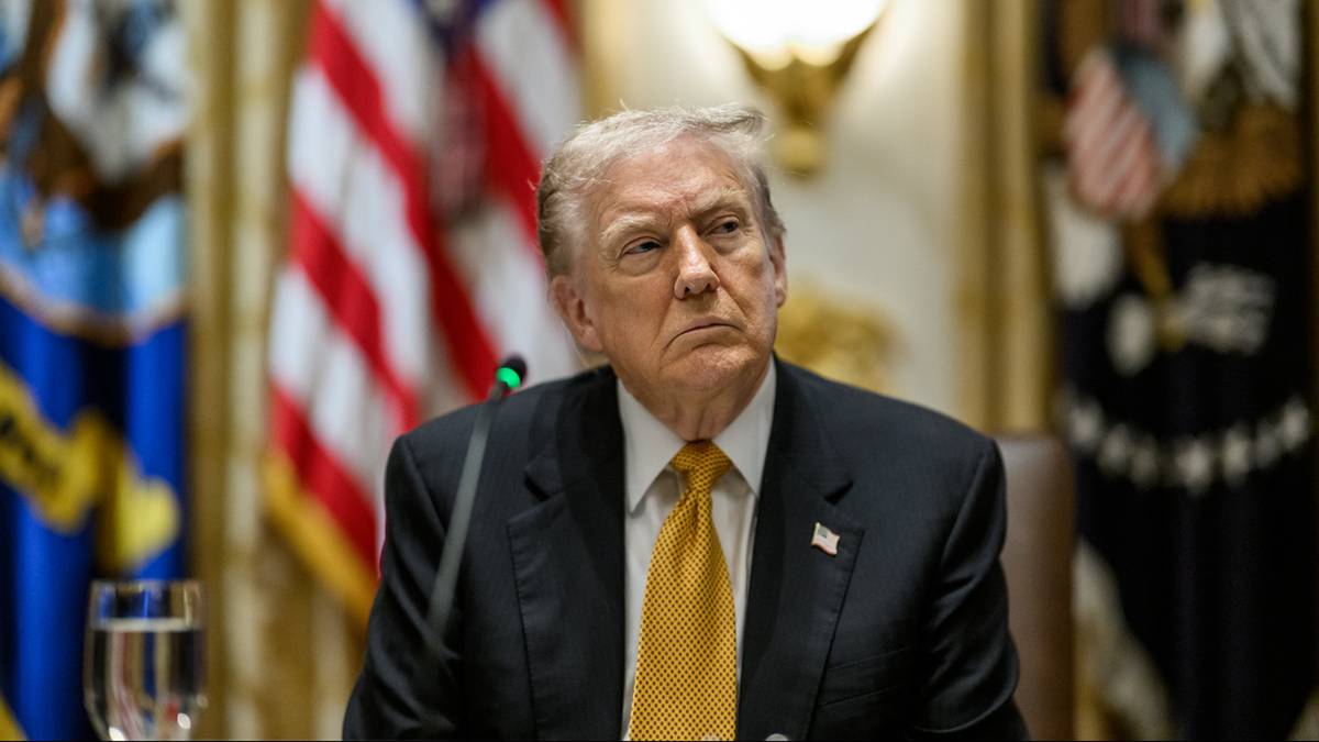 Donald Trump w garniturze i żółtym krawacie, siedzący przed flagami USA.