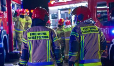 Dwóch strażaków w hełmach i odblaskowych kurtkach z napisem "PAŃSTWOWA STRAŻ POŻARNA" stoją plecami do widza, obserwując wozy strażackie w nocy.