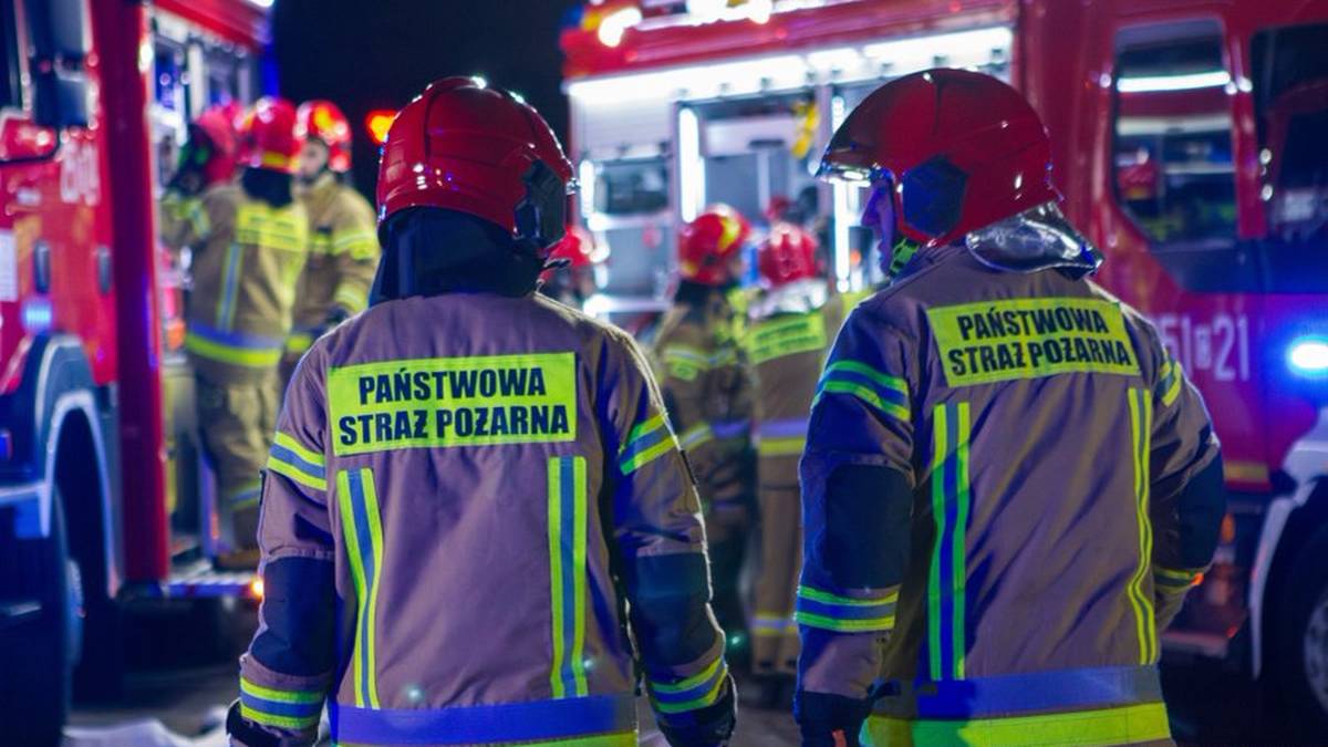 Dwóch strażaków w hełmach i odblaskowych kurtkach z napisem "PAŃSTWOWA STRAŻ POŻARNA" stoją plecami do widza, obserwując wozy strażackie w nocy.