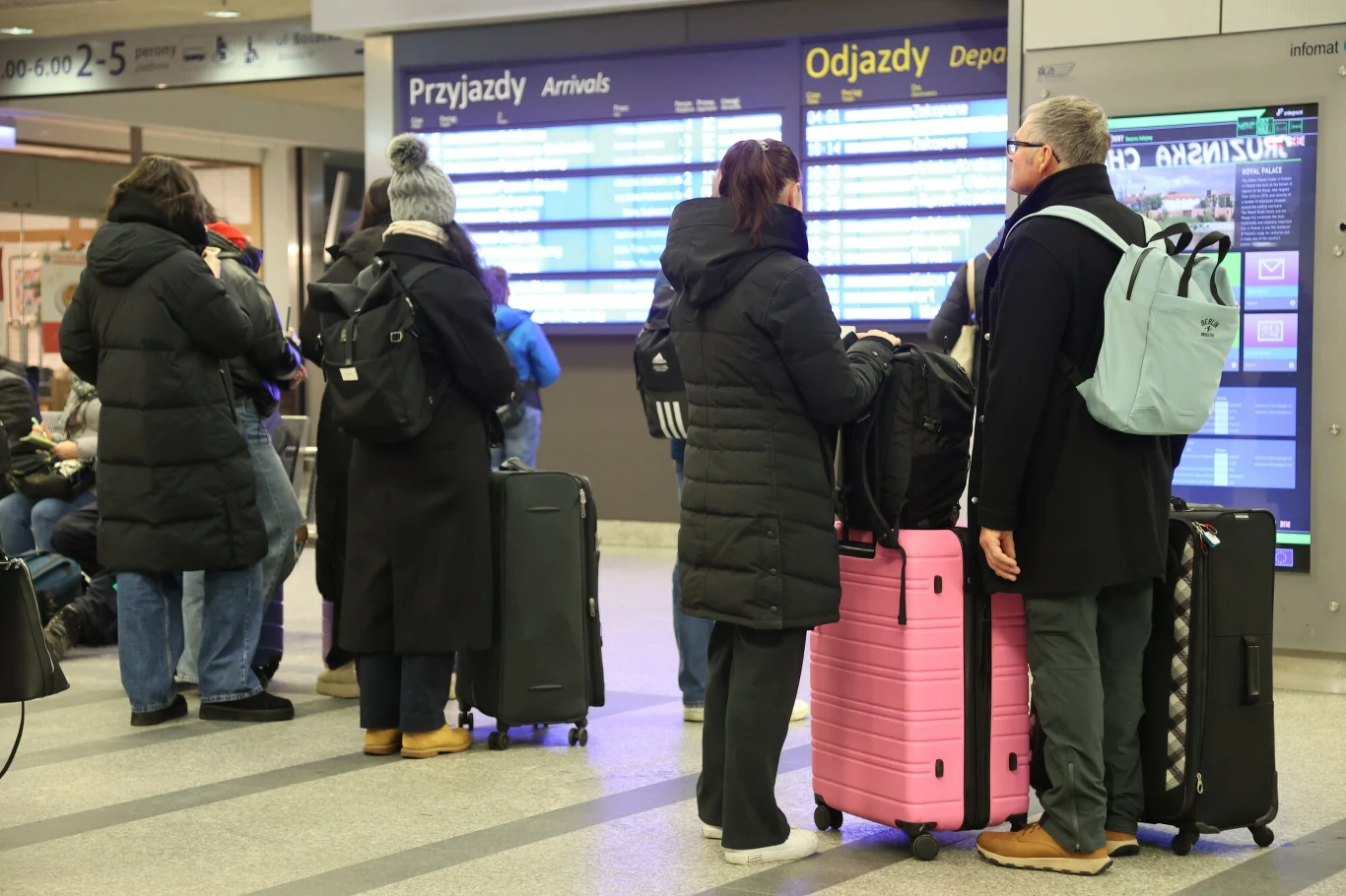 Pasażerowie z walizkami stoją na dworcu, czekają na pociąg przy tablicy z rozkładem jazdy 