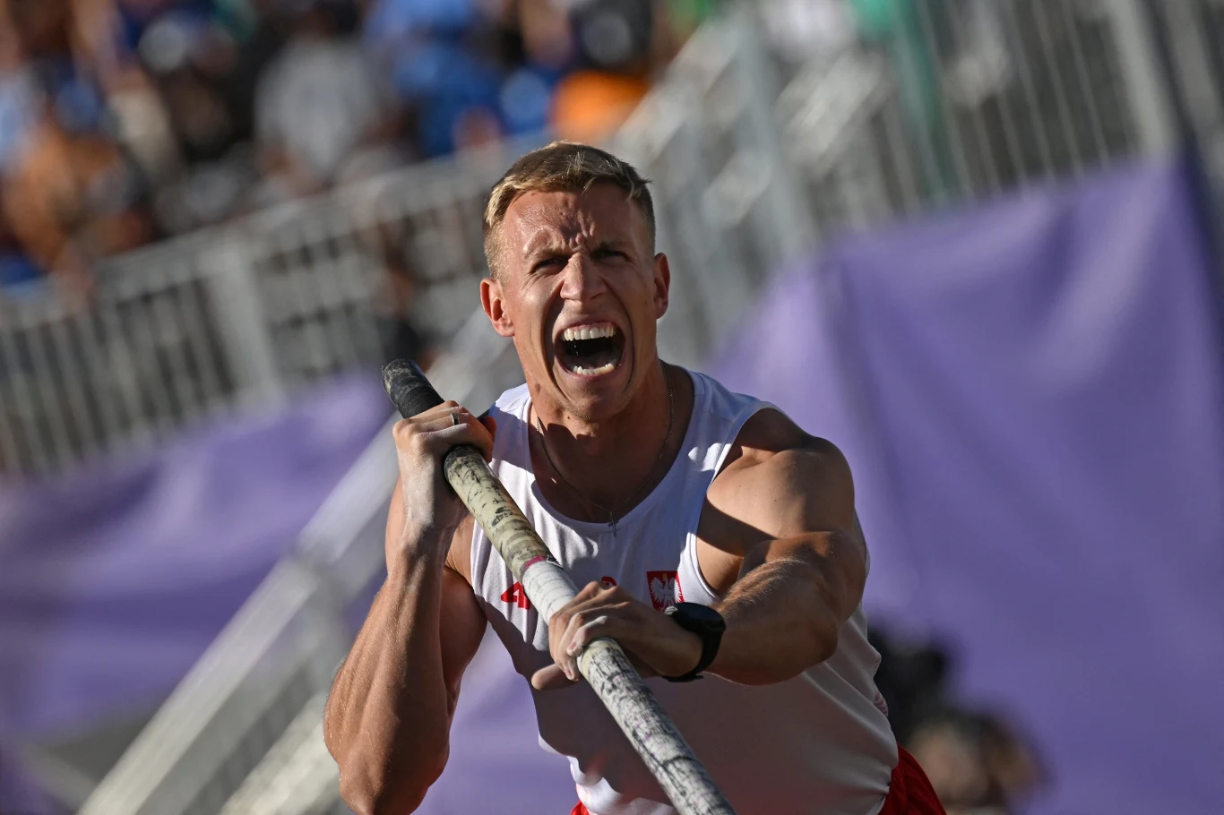 Zawodnik w stroju sportowym z intensywnym wyrazem twarzy trzymający tyczkę podczas skoku o tyczce, na trybunach w tle rozmazana publiczność