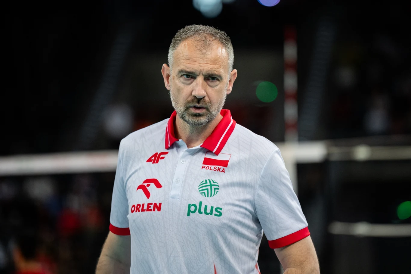 Trener reprezentacji Polski Nikola Grbić dorosły mężczyzna w sportowej koszulce z logami sponsorów oraz naszywką z napisem Polska, stojący na tle niewyraźnego obiektu sportowego