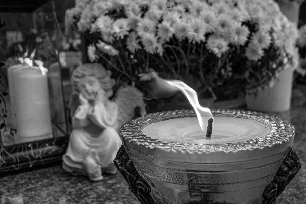 Płonący znicz ustawiony przed figurką aniołka i bukietem białych kwiatów, kompozycja utrzymana w tonacji czarno-białej odwołująca się do zadumy i pamięci o zmarłych.
