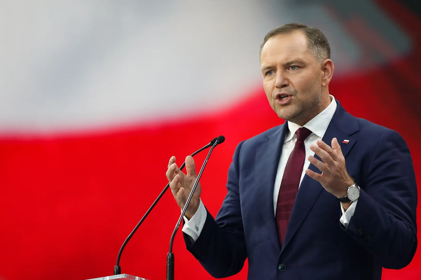 Mężczyzna w garniturze przemawia zza mównicy gestykulując dłońmi na tle rozmytej biało-czerwonej flagi Polski.